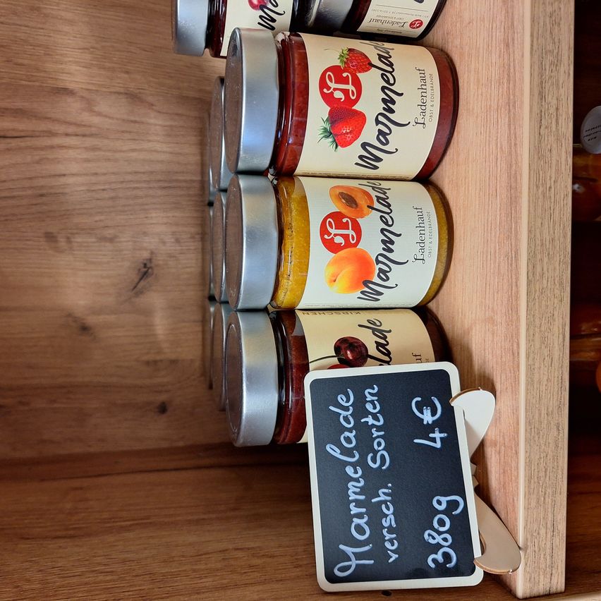 Ein Holzregal zeigt fünf Gläser mit hausgemachter Marmelade in verschiedenen Geschmacksrichtungen, jedes mit einem silbernen Deckel und einem kleinen Preisschild aus Kreide.