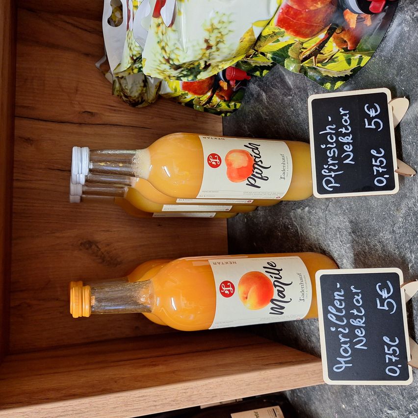 Zwei Flaschen Pfirsichsaft stehen auf einem Holzregal. Eine hat ein Etikett mit der Aufschrift 'Pfirsich' und die andere 'Manille'. Jede Flasche hat einen Preisschild.