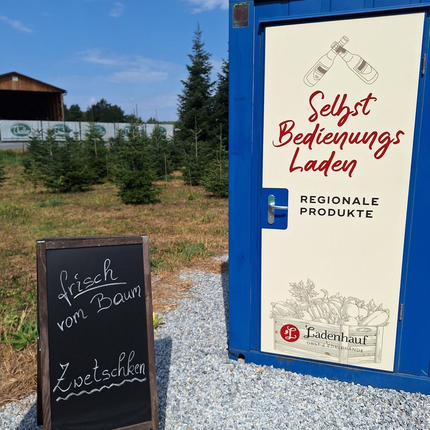 A blue self-service kiosk with a chalkboard sign offering fresh produce from the farm. The kiosk has a door with a handle and a label that reads 'Selfst Bedienungs Laden'.