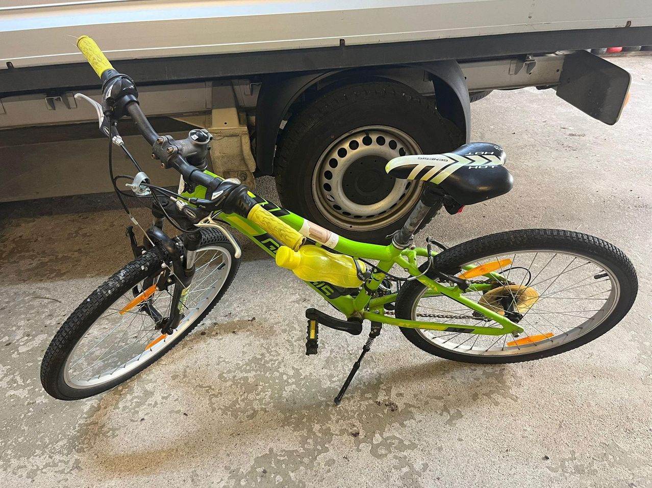 Ein grünes Fahrrad mit einer gelben Wasserflasche ist auf dem Betonboden geparkt. Dahinter steht ein LKW mit einem großen schwarzen Reifen.