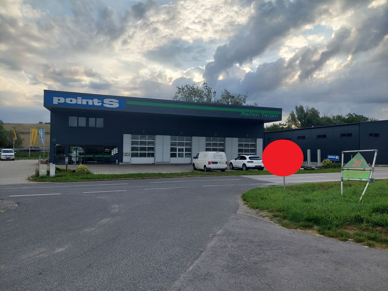Ein Gewerbegebäude mit einem blauen Schild, das 'pointS Reifen Tarjan' steht. Zwei Fahrzeuge sind vor dem Gebäude geparkt, und ein rotes Schild befindet sich rechts. Bäume und Büsche im Hintergrund.