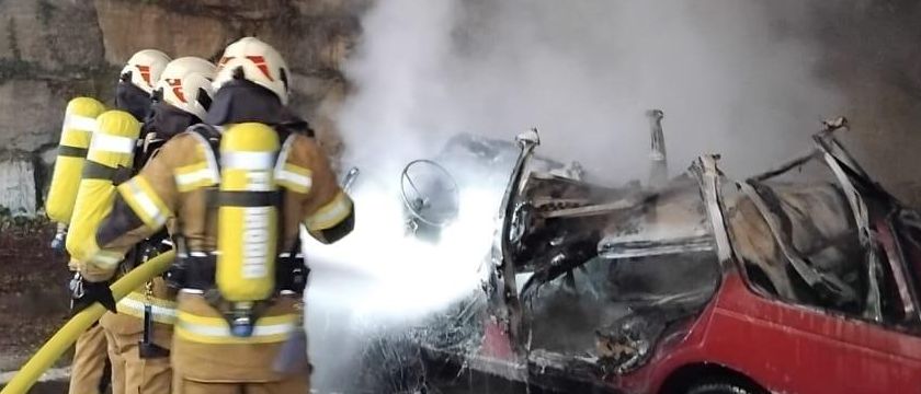 Feuerwehrleute bekämpfen ein Feuer an einem roten Auto. Das Auto steht auf der Straße, stößt Rauch aus, während Feuerwehrleute einen Schlauch benutzen, um das Feuer zu löschen.