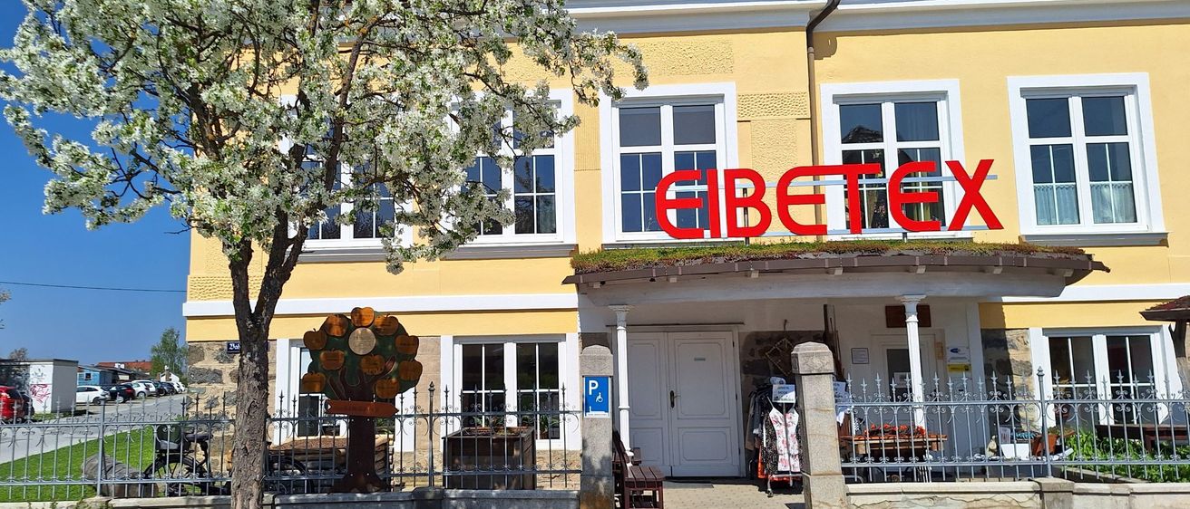 Frontansicht eines gelben Gebäudes mit großen Fenstern, Solarpanels auf dem Dach und einem Schild, das "Eibetex" steht. Das Gebäude hat einen Baum davor und einen Zaun.