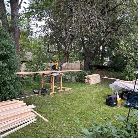 Eine Holzwerkbank in einem Garten, umgeben von Grün. Ein Elektrowerkzeug steht auf der Bank, und in der Nähe befinden sich Holzbretter und ein Trolley. Im Hintergrund stehen Bäume und ein Zaun.