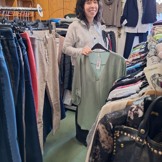 Eine Frau steht in einem Bekleidungsgeschäft, lächelt und hält ein grünes V-Neck-Top in den Händen. Hinter ihr hängen Kleidungsstücke an Kleiderbügeln, und eine Tasche liegt auf dem Boden.