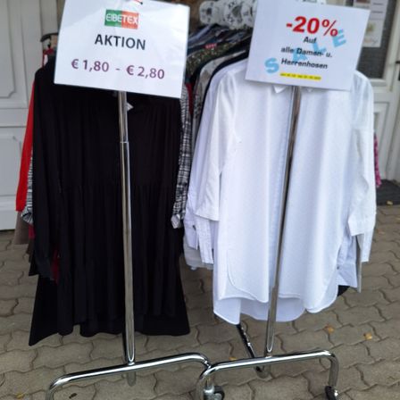 Zwei Kleidungsständer vor einem Geschäft zeigen ein Verkaufsschild mit 20% Rabatt auf alle Damen- und Herrenbekleidung an.