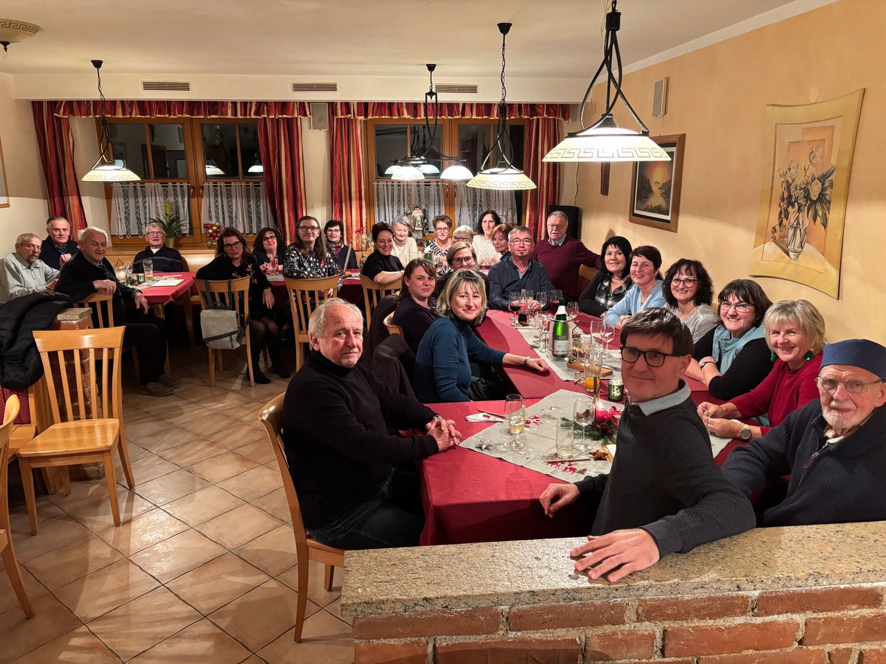 Eine Gruppe von Menschen sitzt an einem Tisch in einem Esszimmer mit roten Tischdecken, Weingläsern und Tellern. Sie lächeln und posieren für ein Foto. Der Raum hat eine Ziegelwand, ein Gemälde und Lampen, die von der Decke hängen.