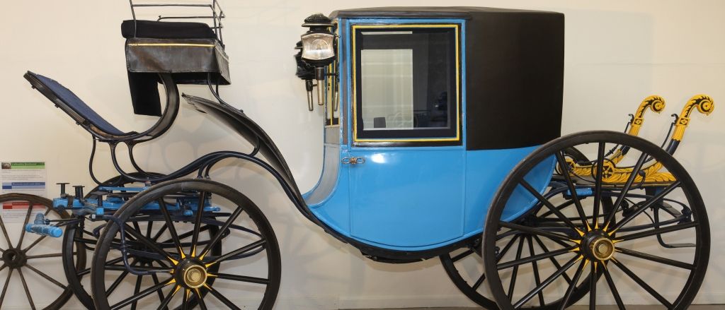 Ein vintage blauer und schwarzer Wagen mit vier Rädern wird auf einem gefliesten Boden ausgestellt. Der Wagen hat ein Glasfenster und eine Lampe, die an der Vorderseite befestigt ist.