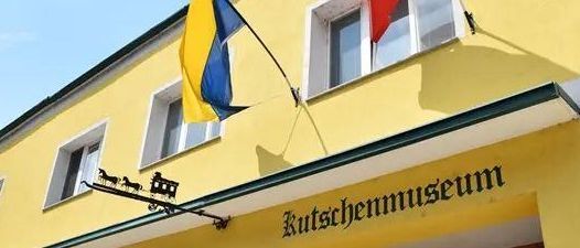 Ein leuchtend gelbes Gebäude mit mehreren Fenstern. Fahnen der Ukraine und Deutschlands hängen am Gebäude. Es gibt ein Schild mit der Aufschrift 'Kutschenmuseum'.