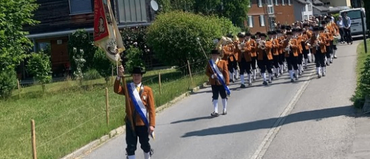 Eine Blaskapelle in traditionellen Uniformen geht mit Flaggen die Straße entlang, umgeben von Grünflächen und Gebäuden.
