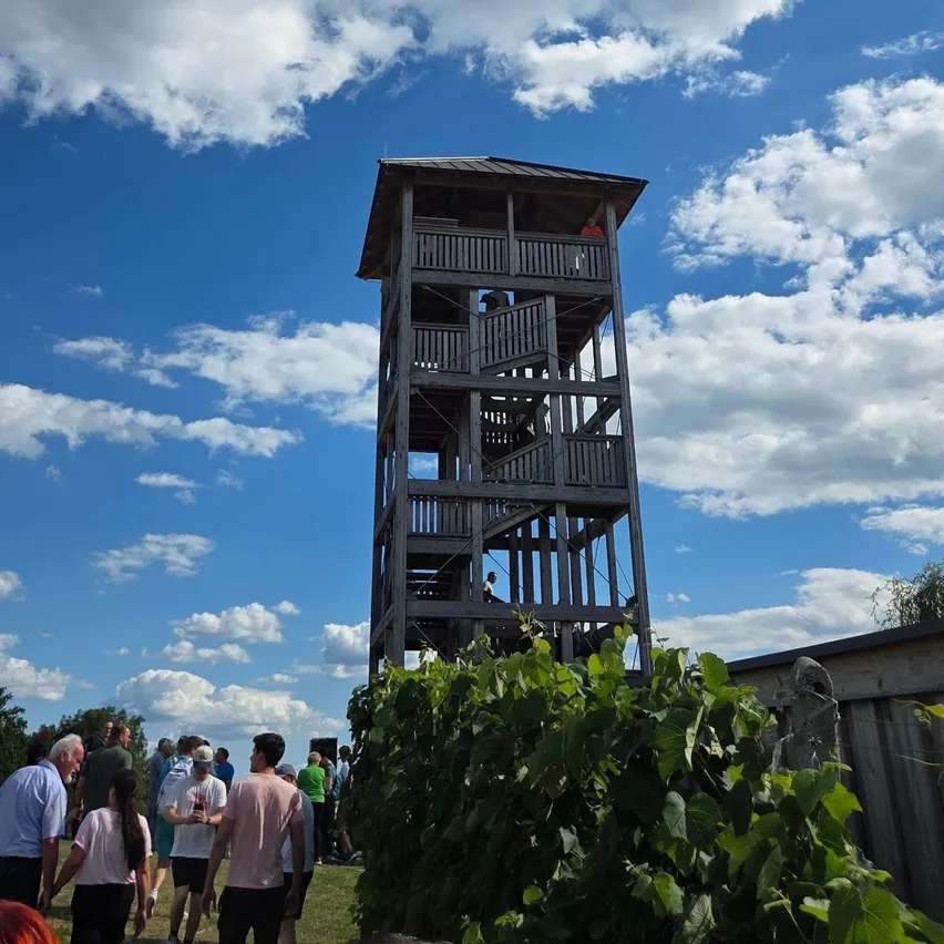 Eine Gruppe von Menschen versammelt sich um einen hohen Holzturm unter einem blauen Himmel mit verstreuten Wolken. Einige Personen steigen die Treppe des Turms hinauf.