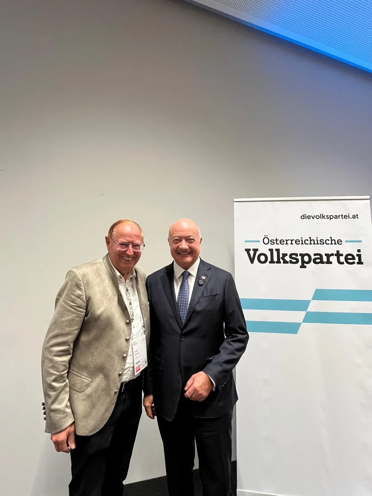 Zwei Männer stehen nebeneinander, lächeln und posieren für ein Foto vor einer weißen Wand mit einem Banner, das 'Österreichische Volkspartei' sagt.