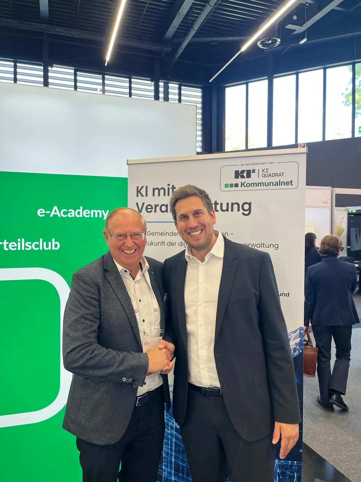 Zwei Männer stehen vor einem grünen Banner mit den Texten 'e-Academy' und 'KI mit Veran'. Sie lächeln und scheinen für ein Foto zu posieren.