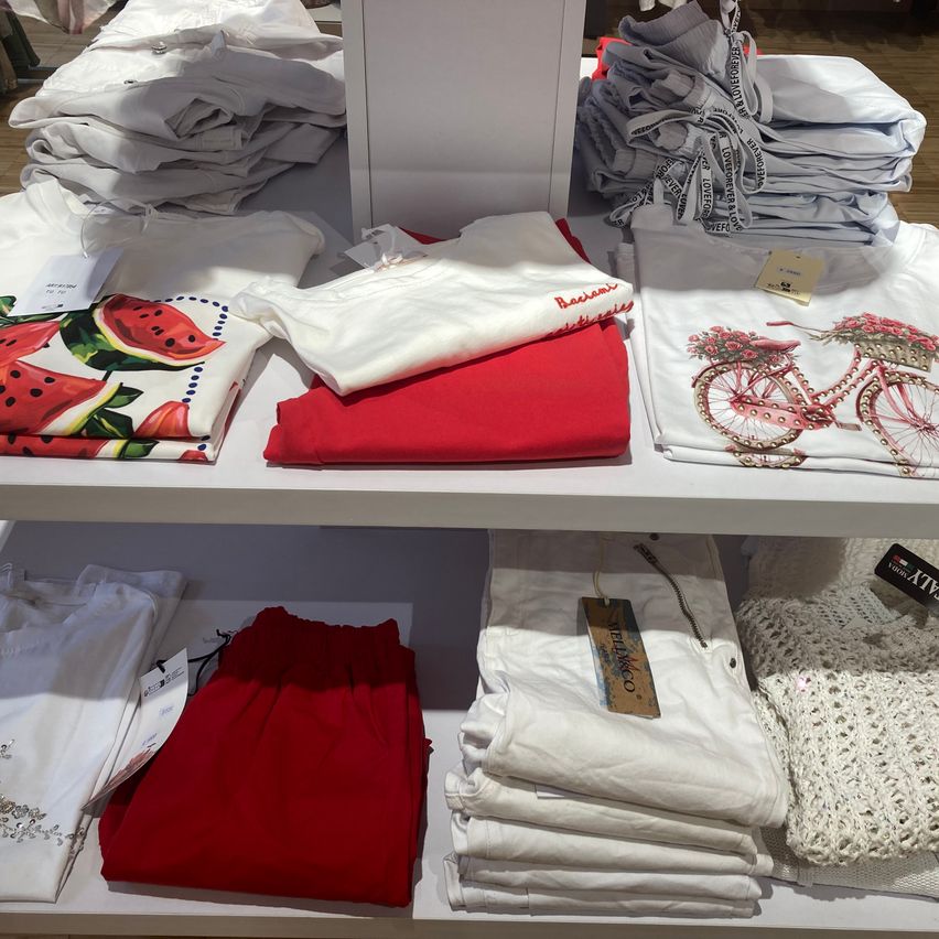 A clothing display features folded shirts in white, red, and floral designs on a white shelf. Some shirts have tags, and there's a crocheted item in white.