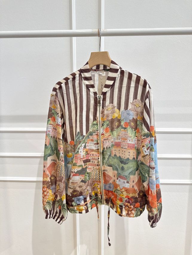 A colorful long-sleeved shirt with a striped pattern and a floral print of a hilly town with buildings and trees is hanging on a white hanger.