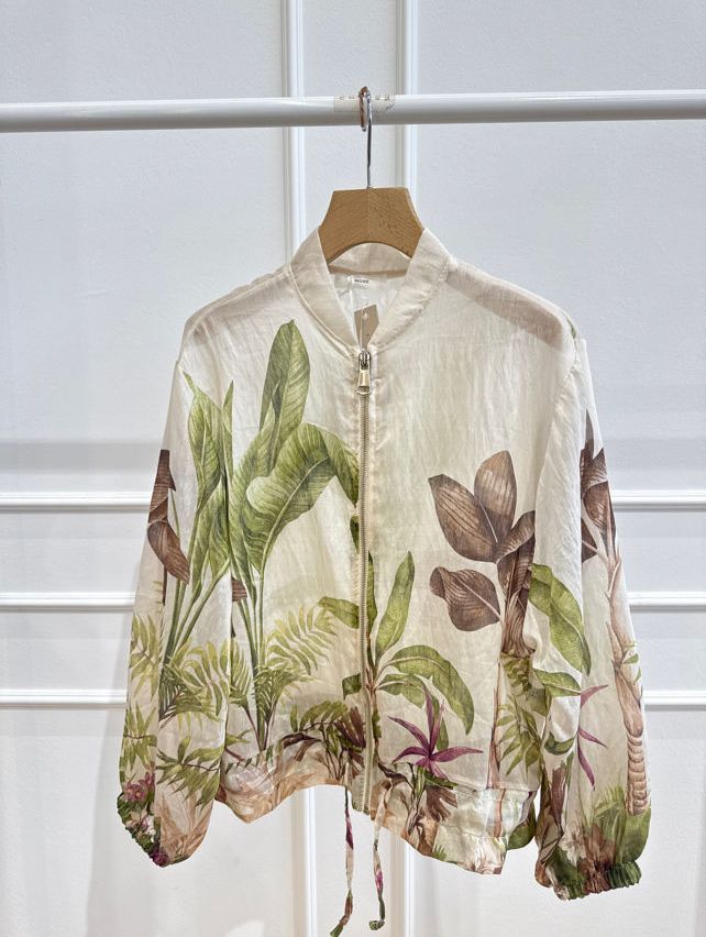 A white jacket with a zipper and a tropical plant print, hanging on a wooden hanger against a white wall.