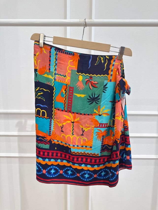 A vibrant, multicolored skirt with a patchwork design featuring tropical elements like palm trees and flamingos is hanging on a wooden hanger against a white background.