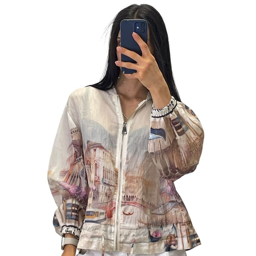 A woman takes a selfie with her phone, wearing a long-sleeve blouse with a printed cityscape design.