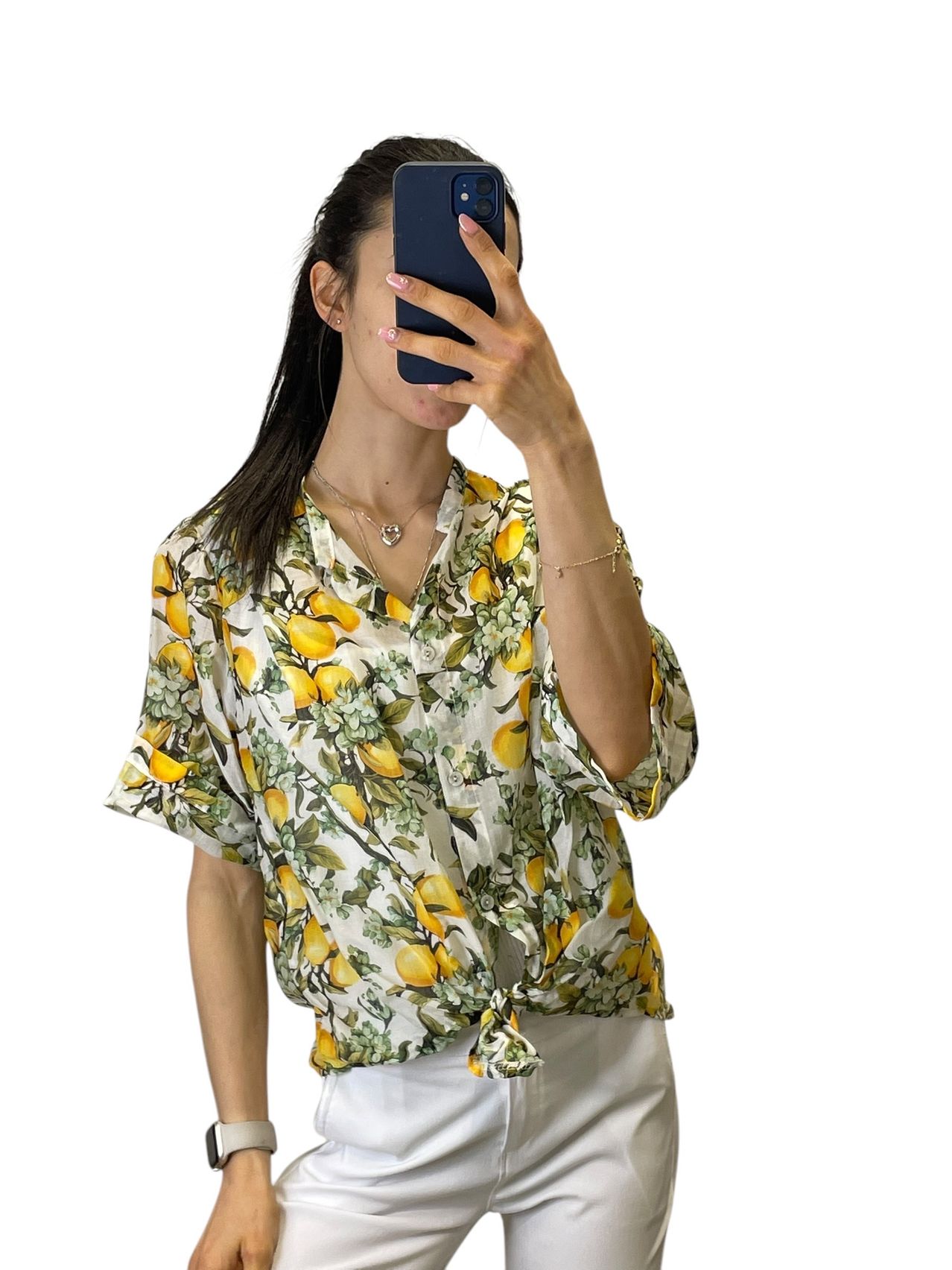A woman takes a selfie with her phone, wearing a white top with a lemon print and white pants. She holds the phone with both hands.