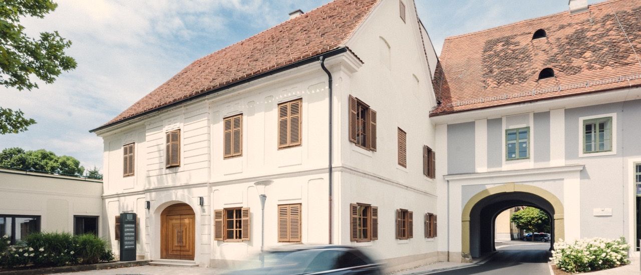 Ein weißes Gebäude mit braunen Fensterläden und einem braunen Dach. Ein schwarzes Auto fährt im Vordergrund vorbei.