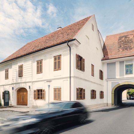 Ein modernes weißes Gebäude mit braunen Fensterläden und einem braunen Dach steht an der Straße. Ein schwarzes Auto fährt schnell auf der Straße.