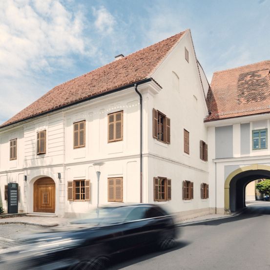 Ein modernes weißes Gebäude mit braunen Fensterläden und einem braunen Dach steht an der Straße. Ein schwarzes Auto fährt schnell auf der Straße.