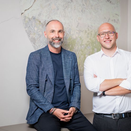 Zwei Männer lächeln und posieren für ein Foto. Sie stehen nebeneinander. Der Mann auf der linken Seite trägt einen blauen Blazer. Der Mann auf der rechten Seite trägt ein weißes Hemd.