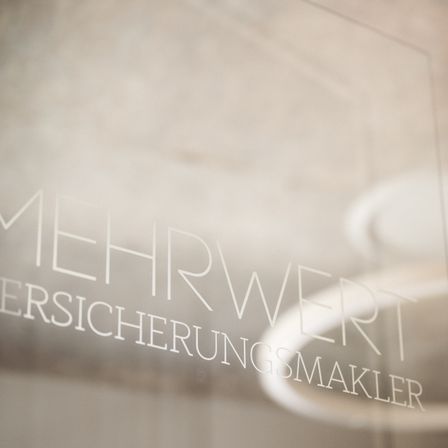 Ein Glaspanel mit weißem Text, der 'MEHRWERT VERSICHERUNGSMAKLER' in einem verschwommenen Hintergrund liest.