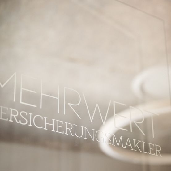 Ein Glaspanel mit weißem Text, der 'MEHRWERT VERSICHERUNGSMAKLER' in einem verschwommenen Hintergrund liest.