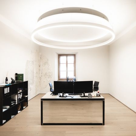 Ein leeres Büro mit einem Schreibtisch und zwei Monitoren, einem Stuhl und einem Bücherregal, beleuchtet von einer kreisförmigen Deckenleuchte. Der Raum hat Holzböden und weiße Wände.