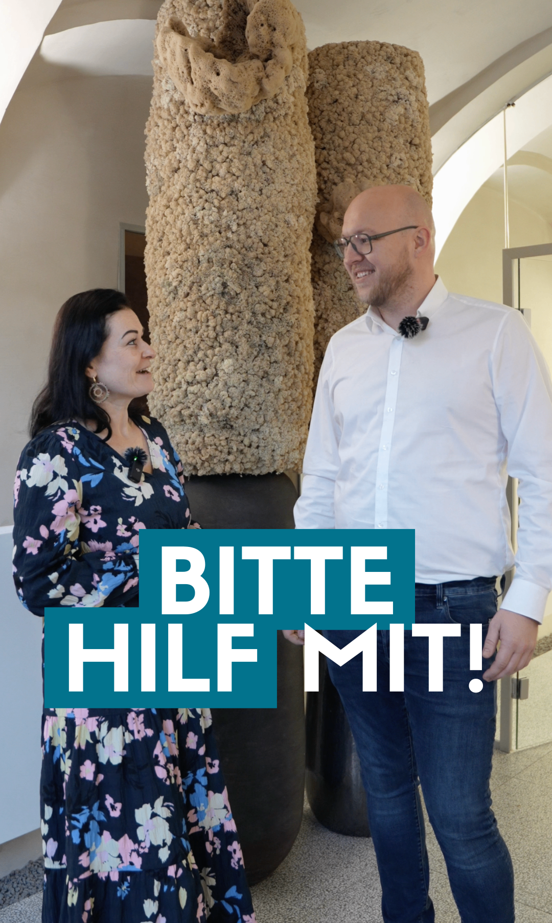 Ein Mann und eine Frau stehen nah beieinander, lächeln und scheinen ein Gespräch zu führen. Die Frau trägt ein Blumenkleid und Ohrringe, während der Mann ein weißes Hemd und Jeans trägt. Hinter ihnen befindet sich eine strukturierte, beigefarbene Wand.