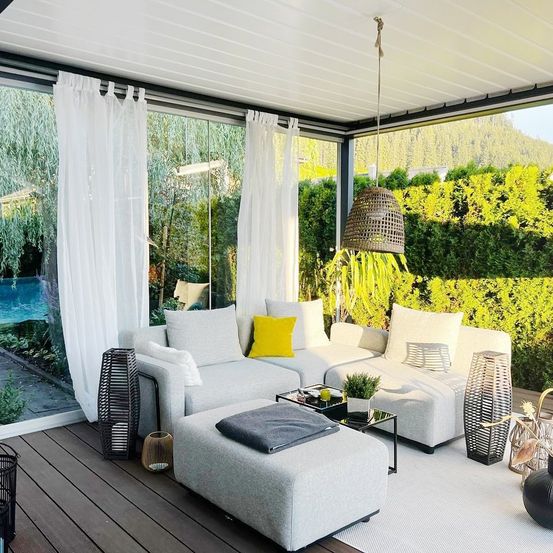 Ein moderner Außenbereich mit einer Glaswand, weißen Vorhängen und einem grauen Sofa. Es gibt ein gelbes Kissen, einen Couchtisch und eine Lampe, die an der Decke hängt.