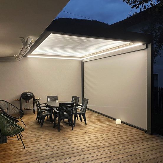 Eine moderne Terrasse bei Nacht mit aufrollbarem Dach und Deckenleuchten. Ein Esstisch und Stühle sind aufgebaut, ein Grill ist in der Nähe.
