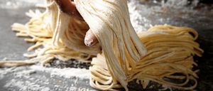 Eine Hand hält einen Bund frischer Pasta-Nudeln, die mit Mehl bestreut sind, in einer rustikalen Küchenumgebung.