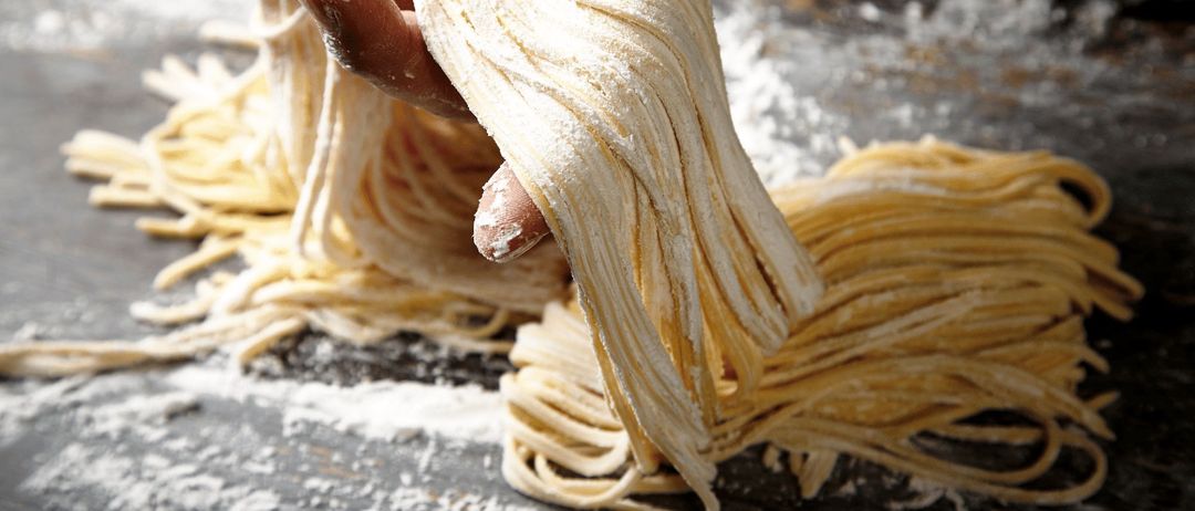 Eine Hand hält einen Bund frischer Pasta-Nudeln, die mit Mehl bestreut sind, in einer rustikalen Küchenumgebung.