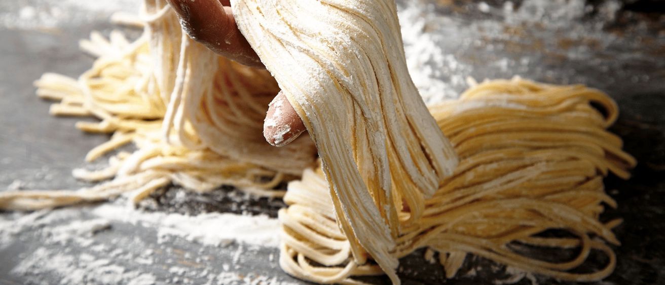 Eine Hand hält einen Bund frischer Pasta-Nudeln, die mit Mehl bestreut sind, in einer rustikalen Küchenumgebung.