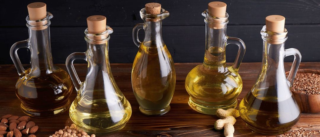 Vier Glasflaschen mit Korkverschluss, gefüllt mit Öl, auf einem Holztisch. Daneben liegen Erdnüsse und Sojabohnen.