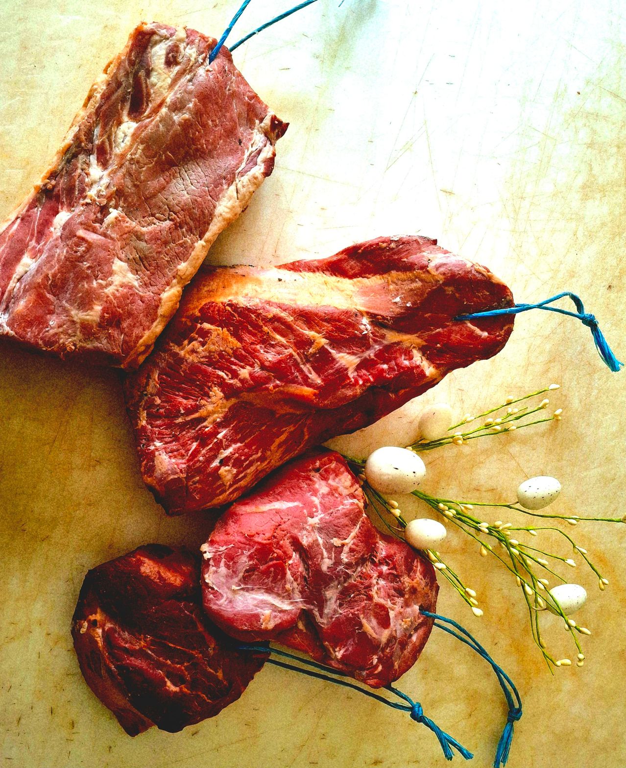 Vier rohe Fleischstücke, die mit blauen Fäden auf einer strukturierten Oberfläche gebunden sind, begleitet von weißen Blumen.