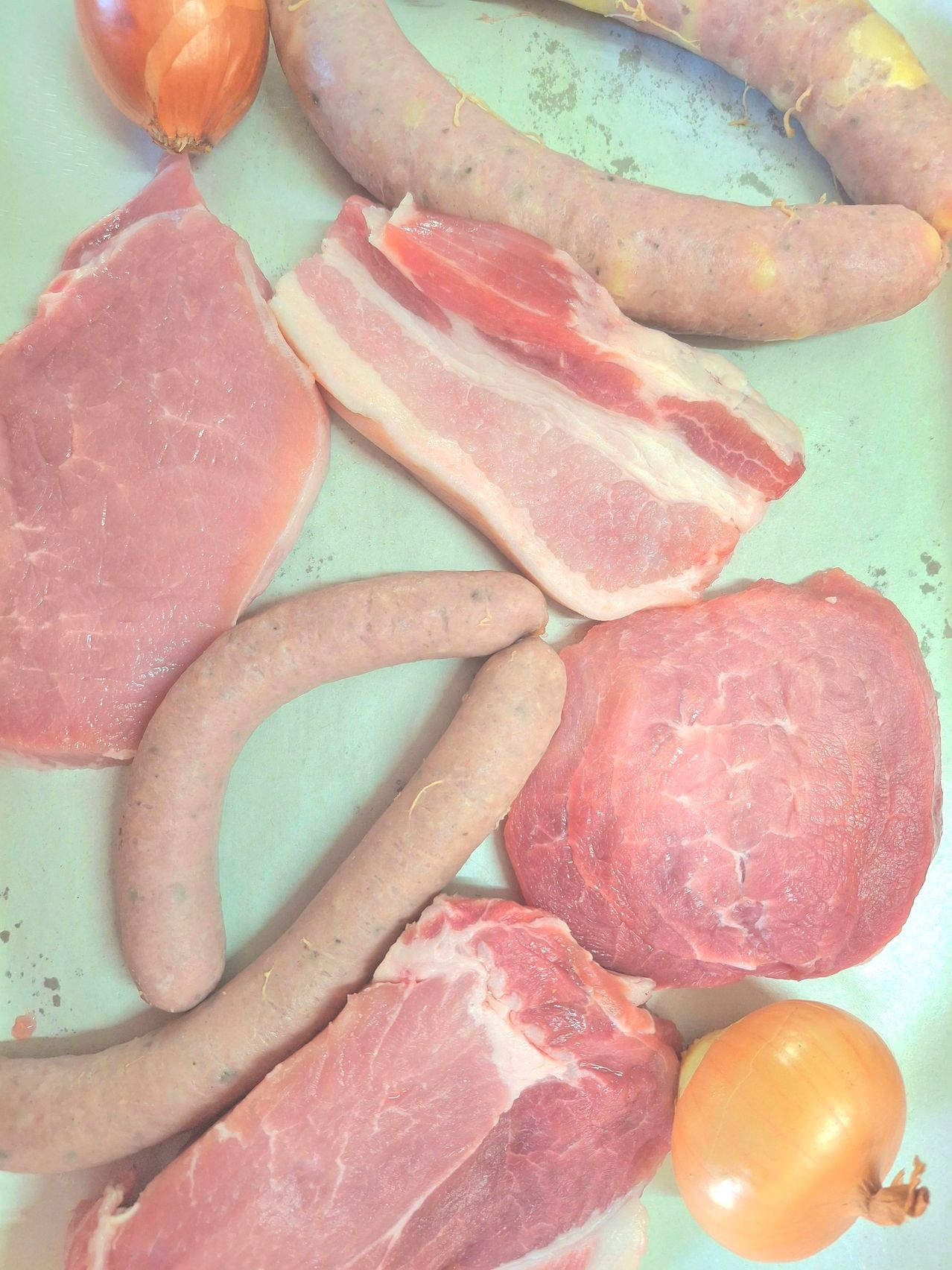 Verschiedene rosa Fleischstücke, einschließlich Wurst und Speck, liegen auf einer grünen Oberfläche. Es gibt auch zwei Stücke Schweinefleisch und eine gelbe Zwiebel.