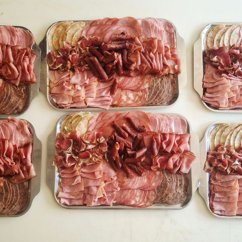 Sechs Metallschalen mit verschiedenen Scheiben von Schinken, Salami und Würstchen, ordentlich auf einer weißen Oberfläche angeordnet. Das Sortiment umfasst verschiedene Arten von Wurstwaren.