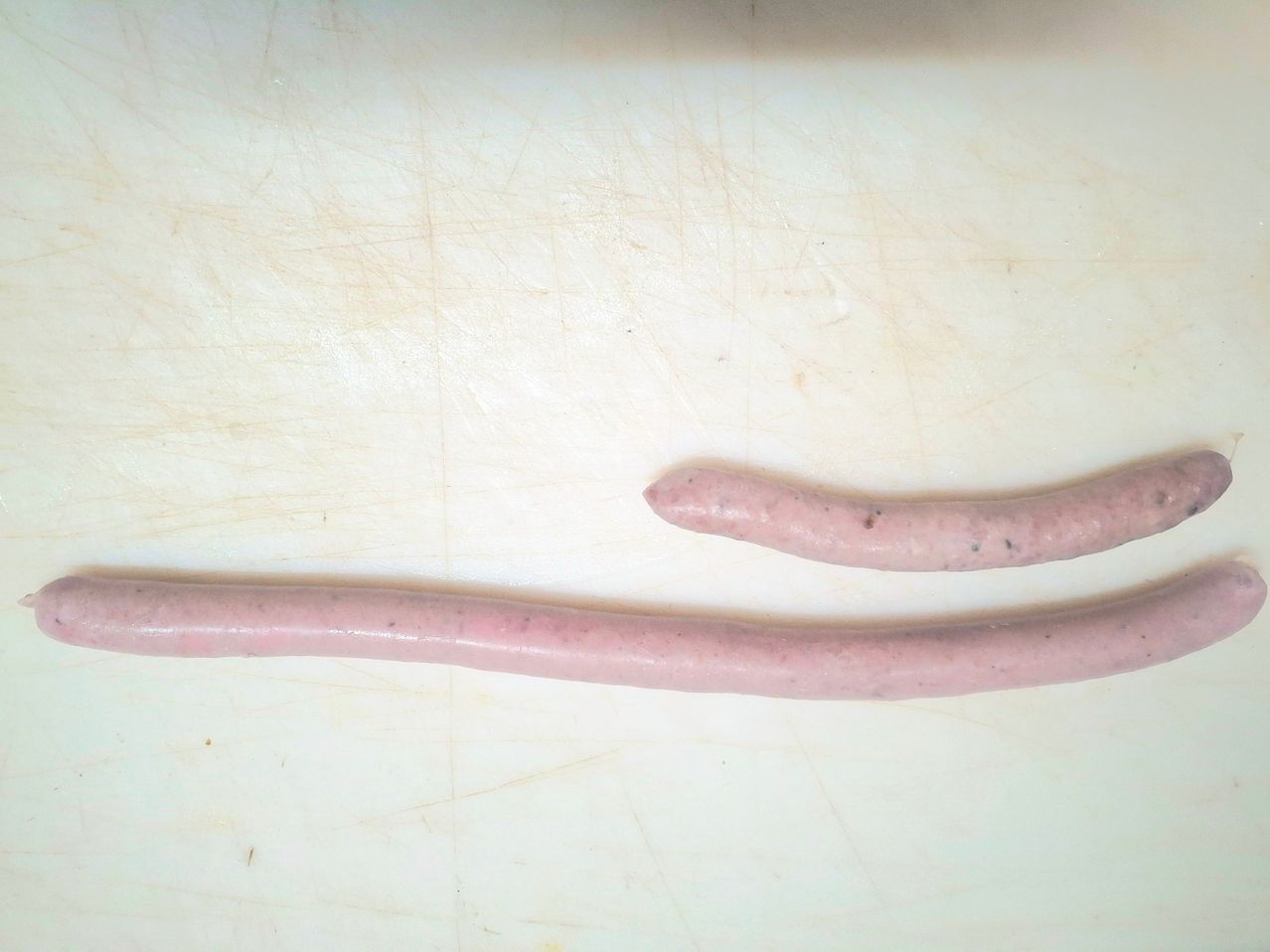 Zwei rosa Würste auf einem weißen Schneidebrett. Ein Wurst ist länger und dünner.
