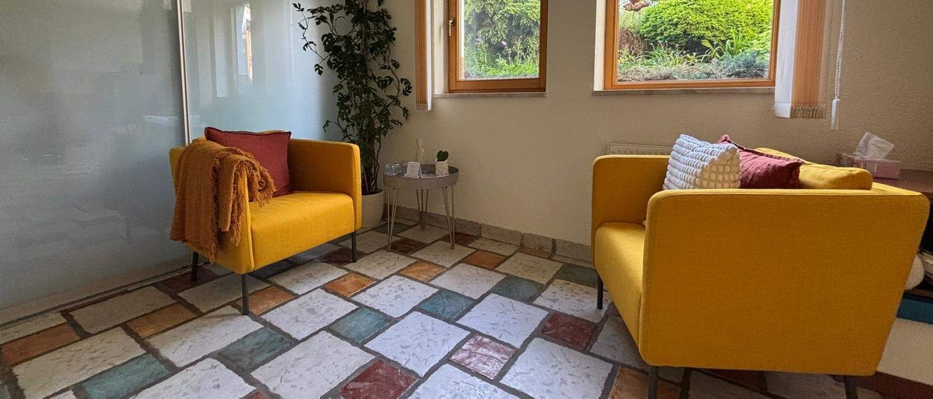 Ein Zimmer mit zwei gelben Sesseln, einem farbenfrohen Fliesenboden und zwei Fenstern. Es gibt eine Pflanze zur Seite und einen kleinen Tisch mit Pflanzen.