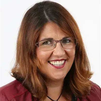 Eine Frau mit braunen Haaren, die eine Brille, eine kastanienbraune Jacke und ein schwarz-weißes Blumenhemd trägt. Sie lächelt.