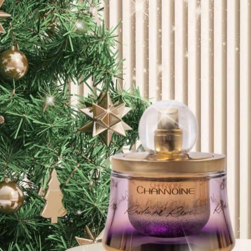 Eine Flasche Channoine-Parfum mit goldenem Deckel, für Weihnachten dekoriert, vor einem geschmückten Weihnachtsbaum platziert.