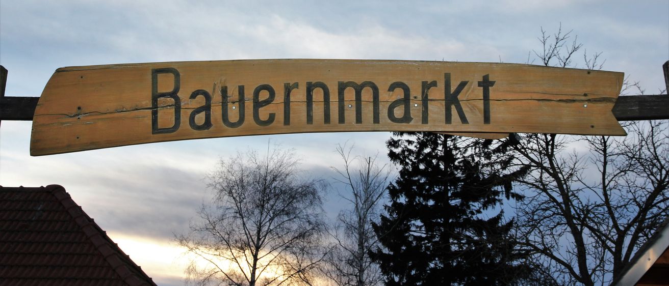 Ein hölzernes Schild mit der Aufschrift 'Bauernmarkt' hängt über einem Dach mit Bäumen im Hintergrund unter einem bewölkten Himmel.