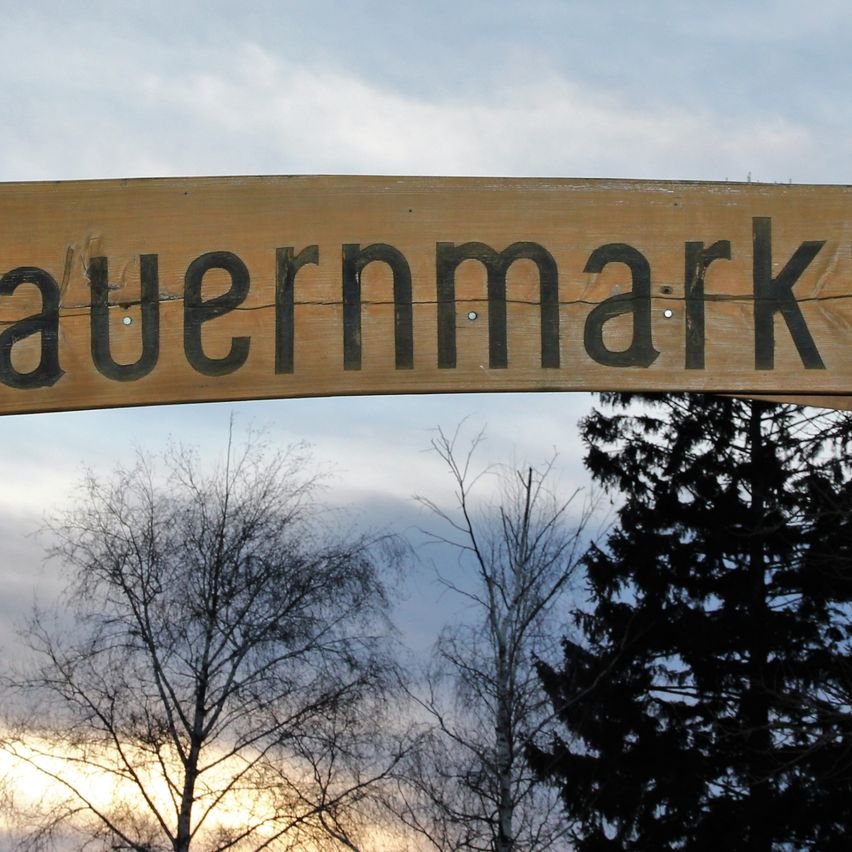 Ein hölzernes Schild mit der Aufschrift 'Bauernmarkt' hängt über einem Dach mit Bäumen im Hintergrund unter einem bewölkten Himmel.