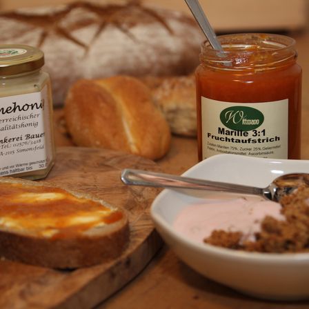 Ein Schneidebrett aus Holz mit einem Stück Brot und einem Glas Marmelade. Ein Löffel ist in einer Schüssel mit Müsli. Dahinter befinden sich zwei weitere Gläser Marmelade und ein Laib Brot.