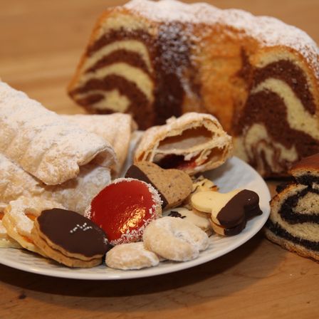 Auf einem Holztisch ist eine Auswahl an Gebäck zu sehen: ein Spiralkuchen, Kekse und ein mit Zuckerguss überzogener Brötchen. Verschiedene Desserts stehen auf einem Teller, darunter ein Schokoladenüberzogener Keks und eine mit Früchten gefüllte Pastete.