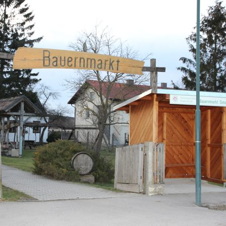 Ein hölzerne Schild steht über einem hölzernen Tor, das zu einem Marktstand führt. Dahinter befindet sich ein Haus mit einem roten Dach, umgeben von Bäumen und Gras.