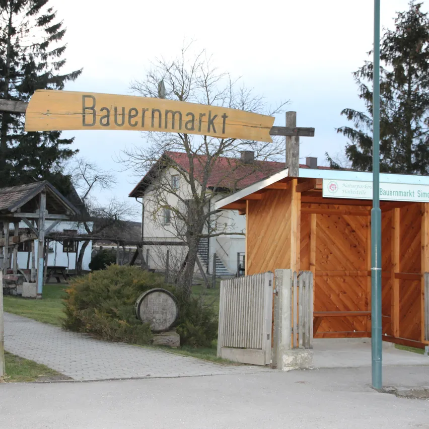 Ein hölzerne Schild steht über einem hölzernen Tor, das zu einem Marktstand führt. Dahinter befindet sich ein Haus mit einem roten Dach, umgeben von Bäumen und Gras.